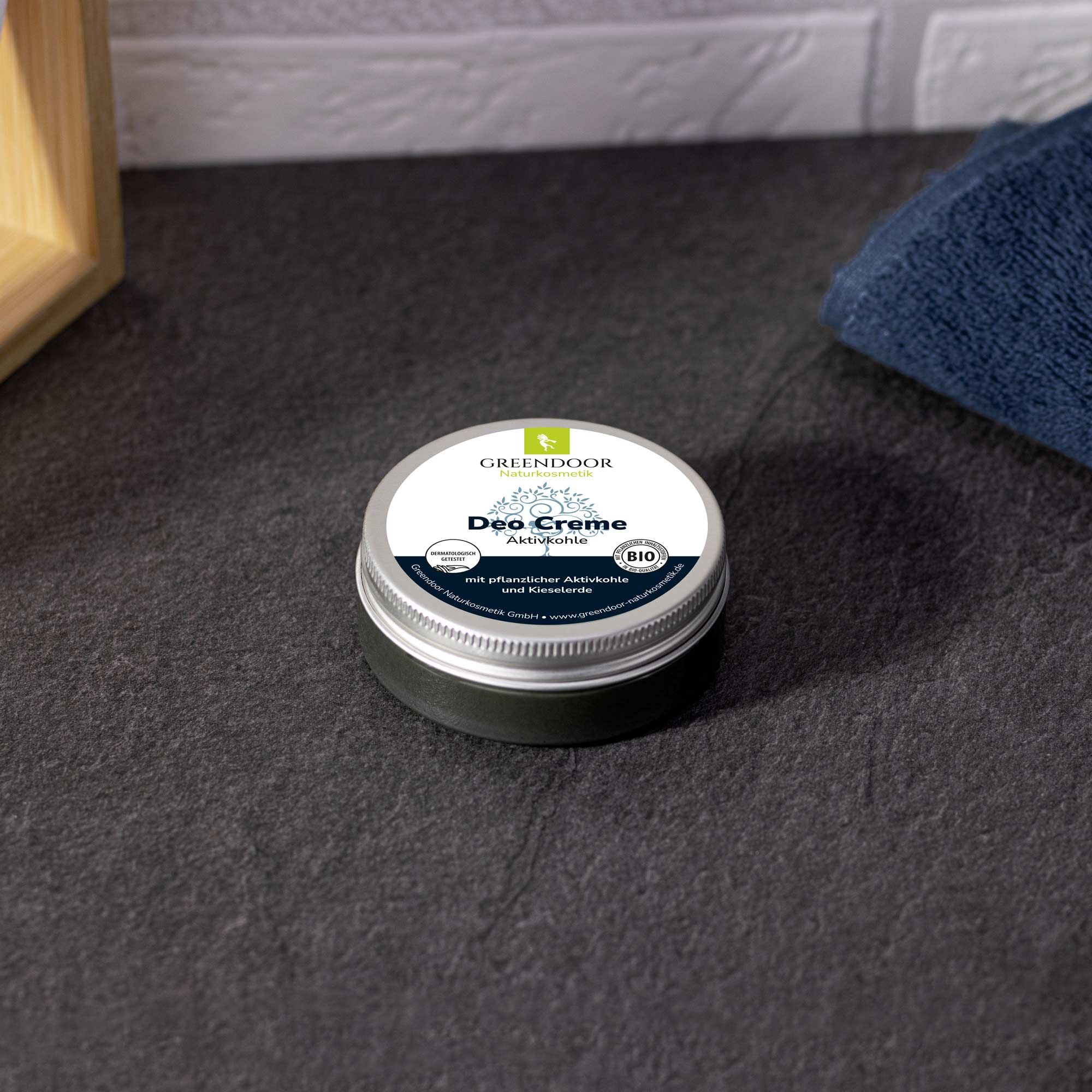 Greendoor Deo Creme mit Aktivkohle auf grauem Untergrund Dose Greendoor Deo Creme mit Aktivkohle, platziert auf dunklem Stein, neben einem blauen Handtuch