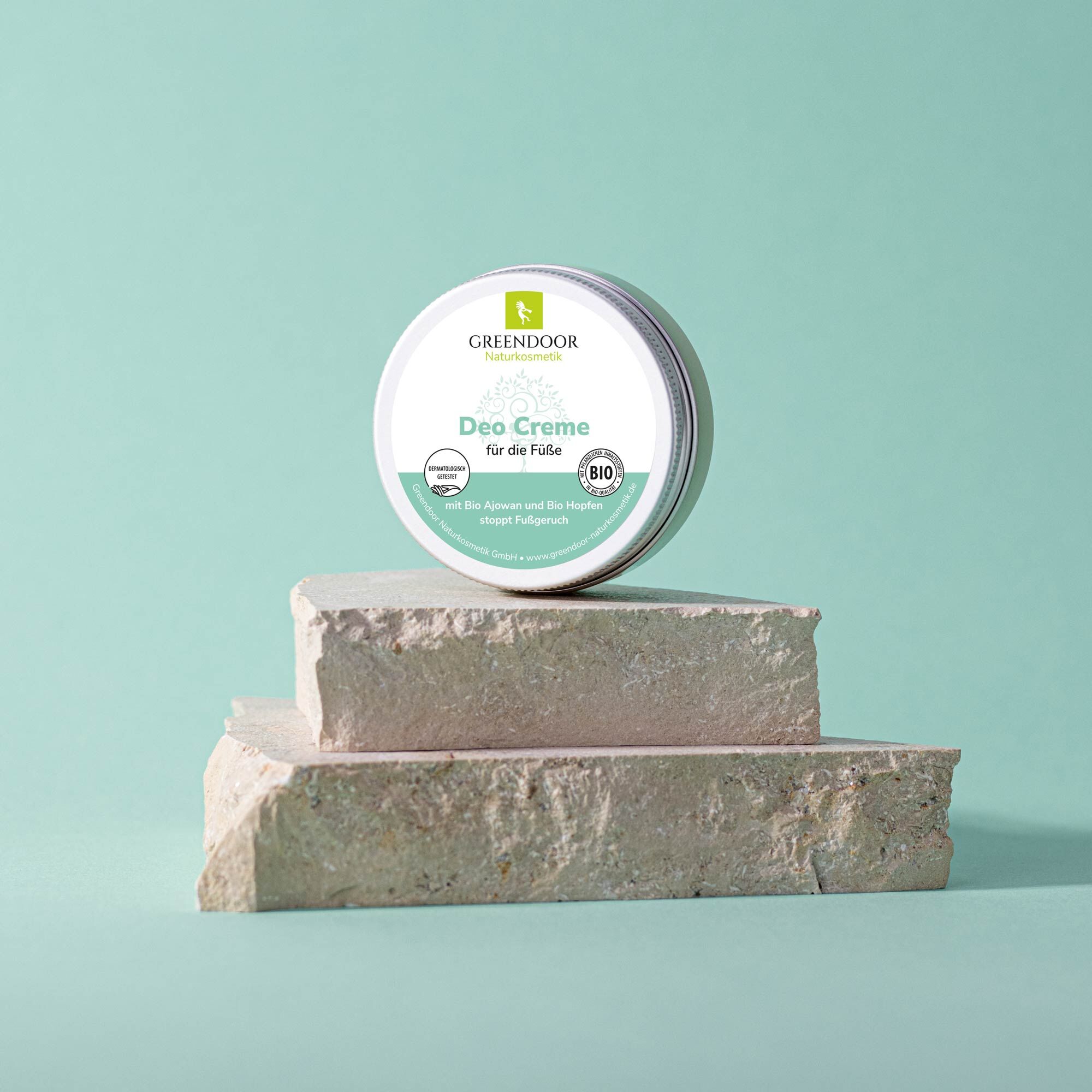 Greendoor Deo Creme für die Füße - Bio Ajowan und Hopfen, stoppt Fußgeruch Eine Dose Greendoor Deo Creme für die Füße steht auf einem Steinpodest vor einem mintgrünen Hintergrund